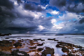 Dramatic stormy sky looms over a rugged ocean coastline with dark foreground rocks.