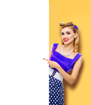 Beautiful smiling woman wear pinup blue dress pointing white blank banner with mockup free text space. Pin up girl advertise, isolated yellow background. Retro vintage sales ad concept