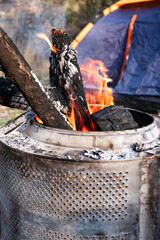 Burning wood logs inside a repurposed washing machine drum used as a diy fire pit at a campsite near a tent