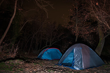 Two camping tents pitched in a dark spooky forest at night surrounded by trees and dry fallen leaves