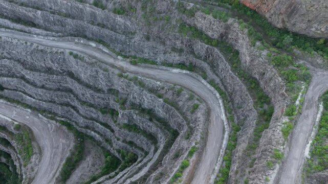 Quarry road bending along terraced rock layers surrounded by sparse vegetation. Mining track curving between steep geological cuts and green patches. Excavation path winding downward through exposed