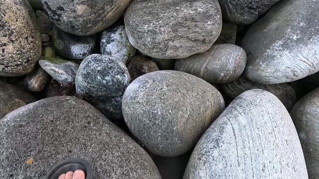 Close walk along a shore of smooth gray stones in Norway, steps in flip-flops tapping over rounded pebbles.