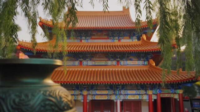 Ancient chinese temple with a traditional bronze incense burner in the foreground. Beautiful historic landmark in asia with lush green willow trees framing the scene