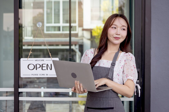 Asian beautiful woman with apron holds laptop, stands next to open sign on glass door at cafe. Young owner starting a small business turning open sign. Happy female waitress working in coffee shop - Powered by Adobe