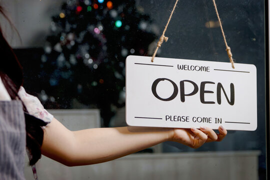Hand of woman hold open sign on glass door at cafe during night shift on Christmas night. Owner starting a small business turning open sign. Female waitress hand with open sign at coffee shop. - Powered by Adobe