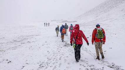 A group of tourists with backpacks are marching along a snowy mountain trail. The view from the back.