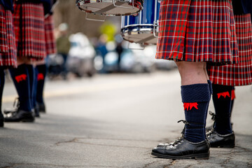 Ground-level detail showing bagpiper's traditional dress including red tartan kilt, black knee socks with red garter flashes, and polished black shoes on pavement.