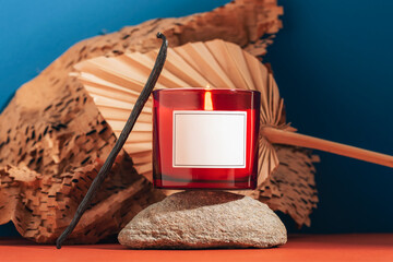Red candle with flame on stone podium with dried palm leaf and textured paper on blue background. Aromatherapy, cozy home atmosphere concept. Closeup