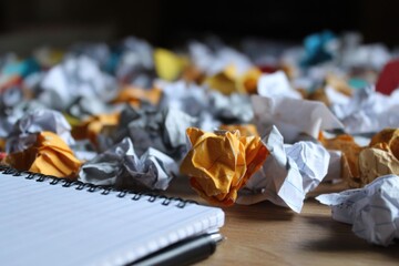 A large number of crumpled papers scattered on a wooden desk beside an open notebook, showing a busy creative process