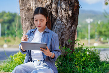 Asian Woman Student Writing on Tablet Device Under Tree Young Female Learning Online Course E Learning Using Pen Studying Outside Education Technology