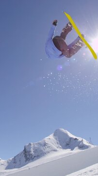 SLOW MOTION: Young pro snowboarder riding the half pipe in big mountain snow park, jumping out of the halfpipe wall and over the sun, performing tricks and rotations with grabs in sunny winter