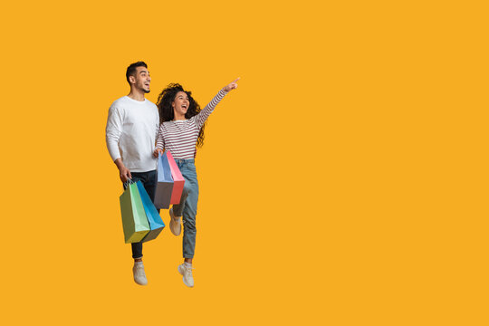 Seasonal Sales Concept. Joyful Arab Couple Jumping With Shopping Bags And Pointing Aside Over Yellow Background, Excited Young Middle Eastern Spouses Enjoying Offers And Discounts, Copy Space