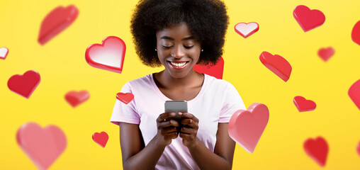 A positive African American woman stands in a yellow studio, using her smartphone. She browses the...