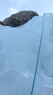POV: Athletic person looking up the mountain while scaling the icy waterfall. Climber carefully picking his spots and placing his ice-picks into the frozen river cascading down the rocky mountain.