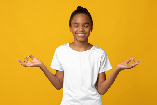 Smiling peaceful cute teen afro american lady pupil in white t-shirt with closed eyes meditation, isolated on yellow background, free space. No stress, happy positive emotions, wellness and lifestyle