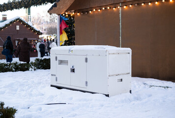 White industrial backup generator in snow at a winter Christmas market with festive lights and wooden stalls.