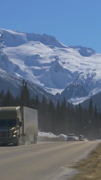 Dust rises from the dirty road as cars and trucks drive down Icefields parkway. Traffic moves along the famous Icefields Parkway highway on a sunny spring day. Snowy mountains overlook a country road