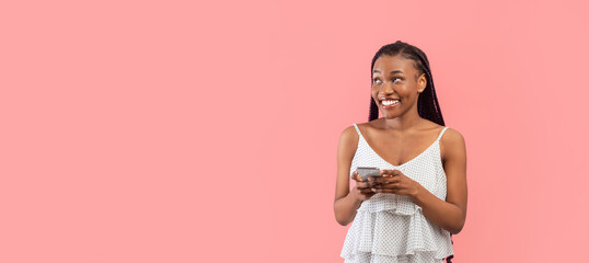 Cheery young black woman using smartphone, chatting online, watching video, looking aside at empty space on pink background, banner design. African American lady surfing web, posting in social media