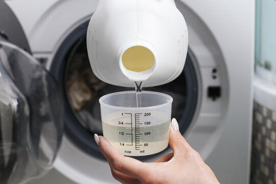 Pouring liquid laundry detergent into washing machine drawer. Measuring cup with washing liquid. Adding detergent before washing cycle. Preparing laundry for washing. Household laundry process.