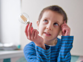 Boy Looking Up in Thought While Holding a Lightbulb for STEM Inspiration - Thoughtful Young Boy Holding a Filament Light Bulb and Scratching His Head