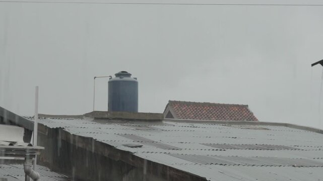 Rain falls over residential rooftops with a water tank visible, capturing a quiet urban scene during overcast weather and everyday life in a calm, natural atmosphere.