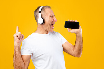 A man stands against a yellow background wearing headphones. He smiles and holds a smartphone while enjoying the music. His expression shows happiness and energy.