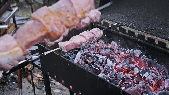 Close-up view of a hand placing metal skewers loaded with marinated meat over glowing charcoal. Smoke rises gently from the black grill emphasizing the start of an outdoor barbecue.