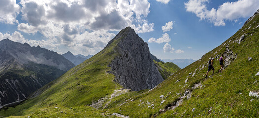 Two hikers ascend a steep trail toward the summit, with another impressive mountain rising in the background at Maldongrat, Hahntennpass, Tyrol, Austria, alpine scenery.