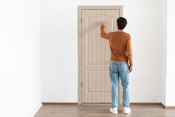 A young man stands at a wooden door, knocking gently with his back turned. He is clothed in a...