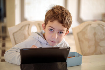 Cute little boy sitting at home table and looking at camera while using digital tablet for remote education or watching cartoons during snack time