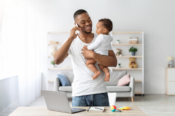 Fototapeta premium Multitasking Concept. Young Black Man Holding Little Baby And Talking On Cellphone While Working Remotely With Laptop At Home, African American Freelancer Guy Enjoying Paternity Leave, Copy Space