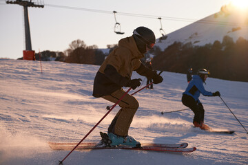 Side view of a skier in a brown suit descending a ski slope at high speed. Alpine skis cut through snow spray at sunset in the Italian Alps with sun rays and blue sky.