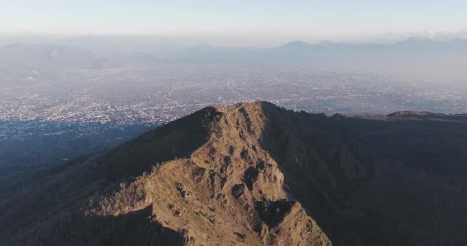 Aerial view of the rugged Mount Vesuvius summit, with its dark, textured slopes contrasting against the distant city haze, Ercolano, Campania, Italy.