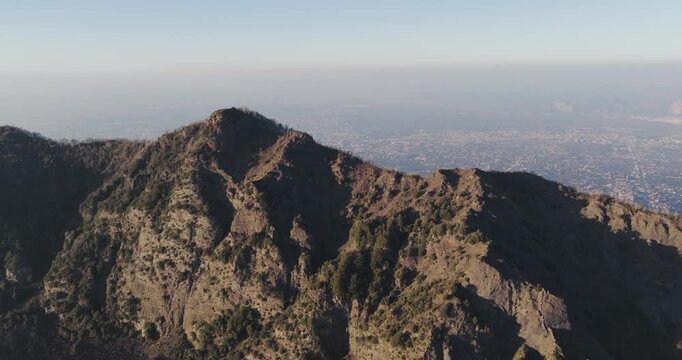 Aerial view of the rugged terrain of Mount Vesuvius, its slopes etched with deep shadows and sparse vegetation, against the backdrop of a hazy urban sprawl, Ercolano, Campania, Italy.