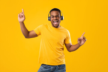 Cheerful Black Guy Singing And Dancing Listening To Music Online Wearing Headphones Having Fun Over Yellow Studio Background. Millennial Man Enjoying Great Playlist. Musical App Concept