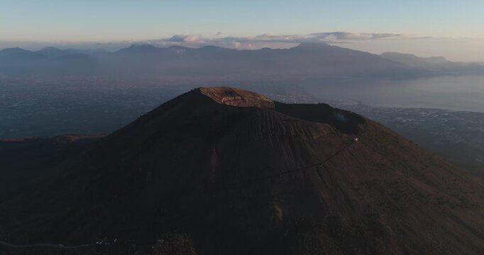 Aerial view of Mount Vesuvius, its dark slopes contrasting with the hazy blue horizon and distant mountains, a serene yet powerful landscape, Ercolano, Campania, Italy.