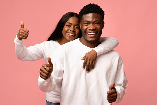 A beautiful black couple is smiling and showing thumbs up while hugging each other in a bright pink studio. Their joyful expressions suggest they are recommending something fun and exciting.