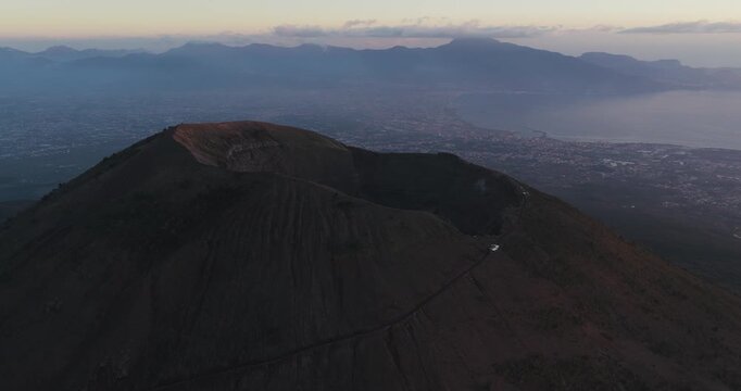Aerial view of Mount Vesuvius's dark, textured crater with a winding path, contrasted against the distant city and sea, Ercolano, Campania, Italy.