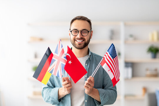 Handsome young guy holding bunch of diverse flags and smiling at camera indoors. Happy millennial man recommending foreign languages school, emigrating abroad. Modern education and student exchange
