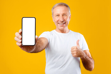 A man stands in front of a yellow background holding a smartphone with a blank screen. He smiles...