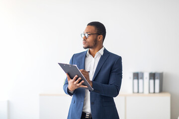 Serious young black psychologist writing in clipboard, looking aside deep in thought at modern office. Psychotherapy services, mental health professional concept