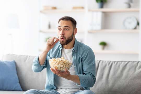 Millennial Caucasian man eating popcorn while watching TV at home. Surprised young guy enjoying interesting program on television, having snack, sitting on couch indoors. Domestic hobbies