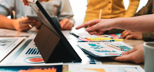 A diverse business team engages in an intense collaborative session, analyzing charts and data on a tablet, surrounded by reports and notes in a modern office environment. SACTR