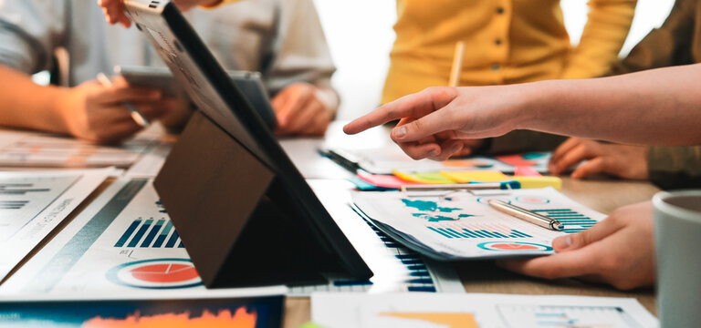 A diverse business team engages in an intense collaborative session, analyzing charts and data on a tablet, surrounded by reports and notes in a modern office environment. SACTR - Powered by Adobe