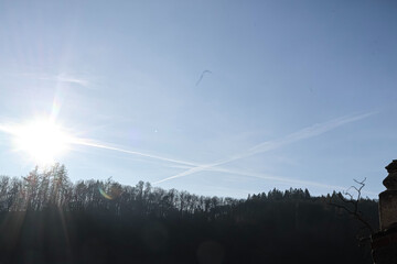Bright sun flares over a dark forest canopy with contrails crossing the sky