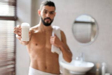 Handsome young guy stands shirtless in a stylish bathroom, holding a tube of deodorant close to the...