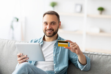 Cheery young guy with credit card and digital tablet sitting on sofa at home, buying goods on web. Attractive Caucasian man using touch pad, shopping on sale in online store during covid quarantine