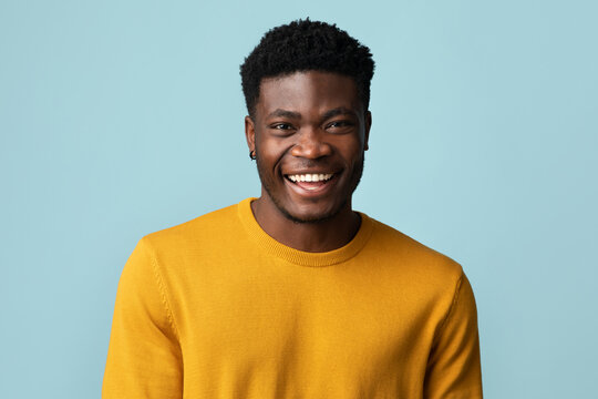Portrait of handsome young black guy in stylish yellow jamper posing alone on blue studio background, cheerfully smiling at camera, copy space. Millennials males guys photos concept