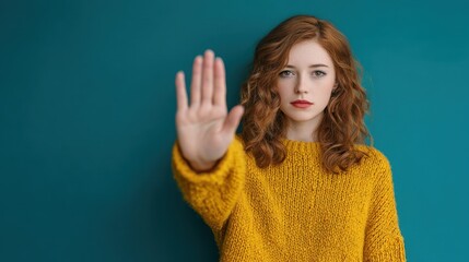 A woman stands against a blue wall. She raises her hand in a stopping gesture while wearing a yellow sweater. The setting appears bright and simple likely indoors during the day.