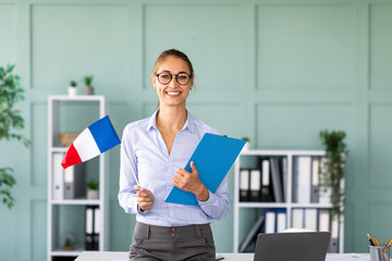 A cheerful teacher stands by her desk, holding a clipboard and the French flag. She smiles at the camera, ready to guide students through their foreign language learning journey.